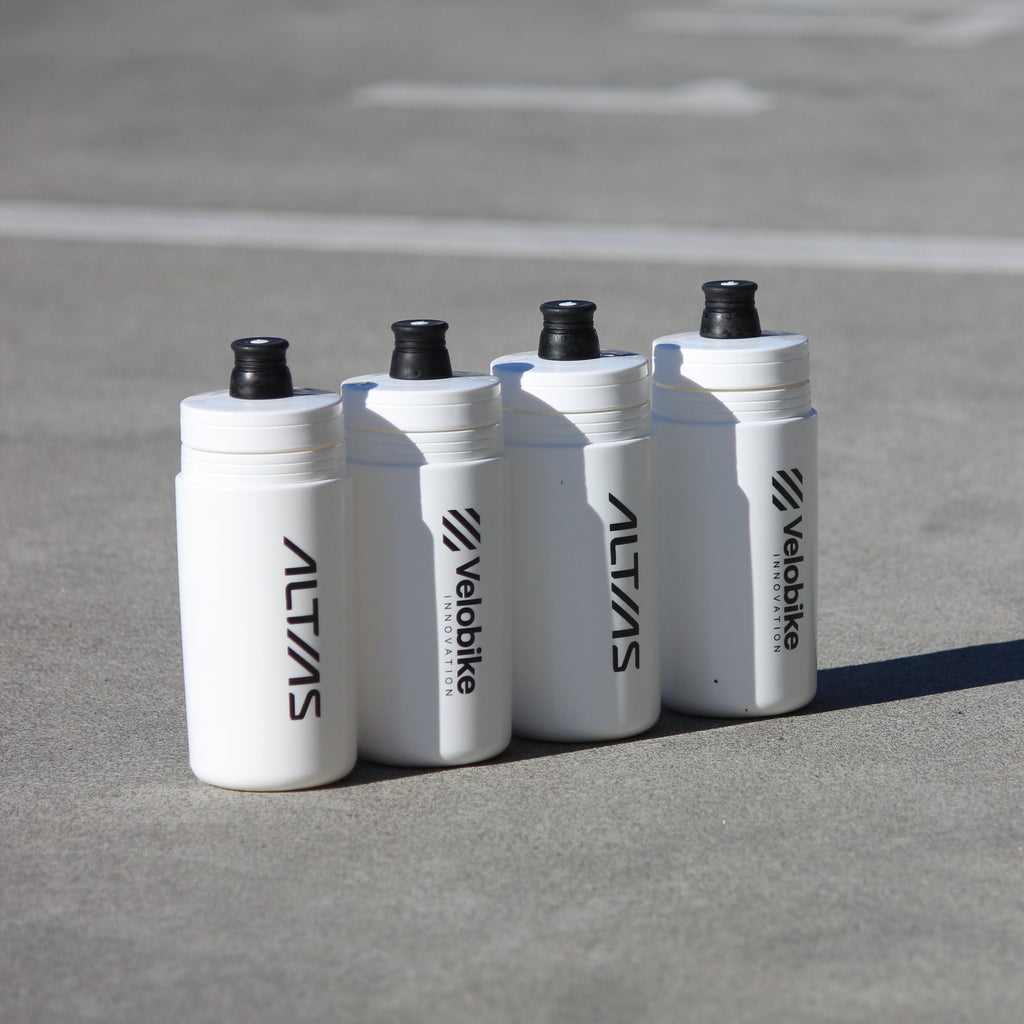 Four white Altias/Velobike water bottles with black caps lined up on a gray concrete surface.