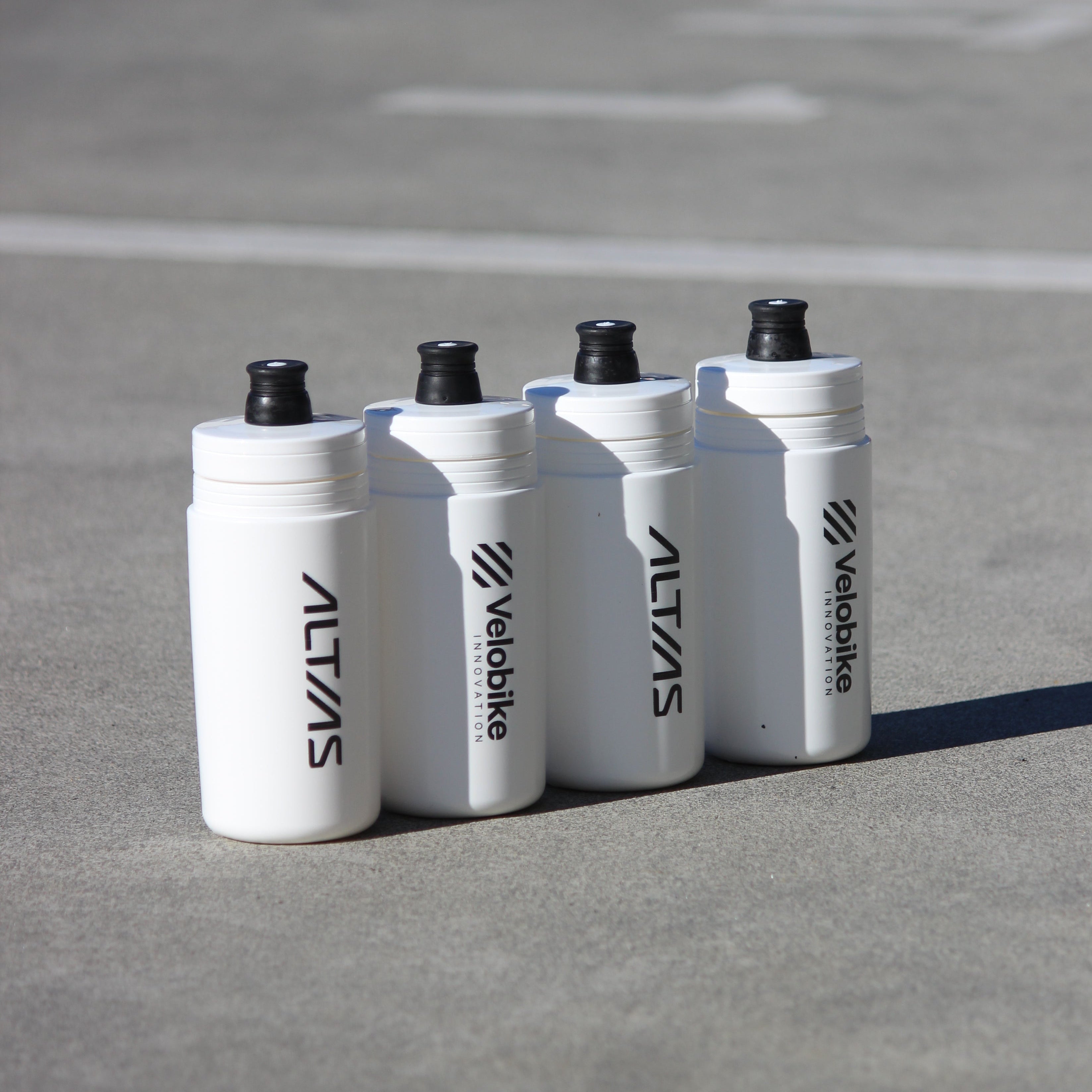 Four white Altias/Velobike water bottles with black caps lined up on a gray concrete surface.