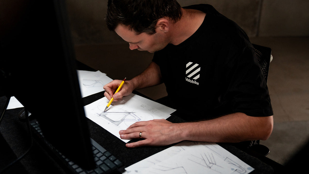 A man wearing a black Off-White t-shirt sits at a desk, drawing on white sheets of paper with a yellow pencil. A laptop is visible in the foreground, and the Off-White brand logo is printed on the chest of the shirt.
